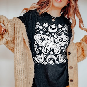Puede incluir: Una camiseta gris oscuro con un gr&aacute;fico blanco de una mariposa rodeada de estrellas, lunas y flores. La mariposa tiene un cuerpo en forma de coraz&oacute;n y alas intrincadas.