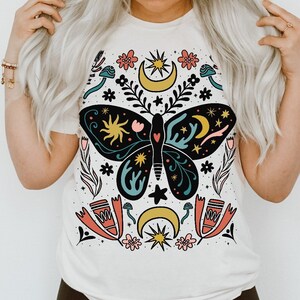 May include: A white t-shirt featuring a colourful butterfly design. The butterfly is surrounded by flowers, stars and moons. The design is in a folk art style.