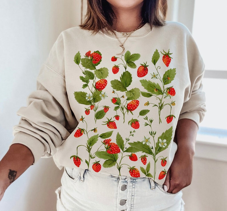 Puede incluir: Sudadera beige con un dise&ntilde;o de acuarela de fresas, hojas verdes y flores blancas. Las fresas son rojas con peque&ntilde;as semillas, y las hojas son de un verde vibrante. El dise&ntilde;o se extiende por la parte delantera de la sudadera.