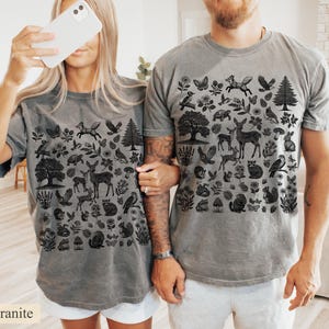 May include: Two gray t-shirts with a black nature-themed design. The design includes illustrations of trees, deer, birds, butterflies, and other woodland elements. The word "granite" is printed at the bottom left corner of the image.