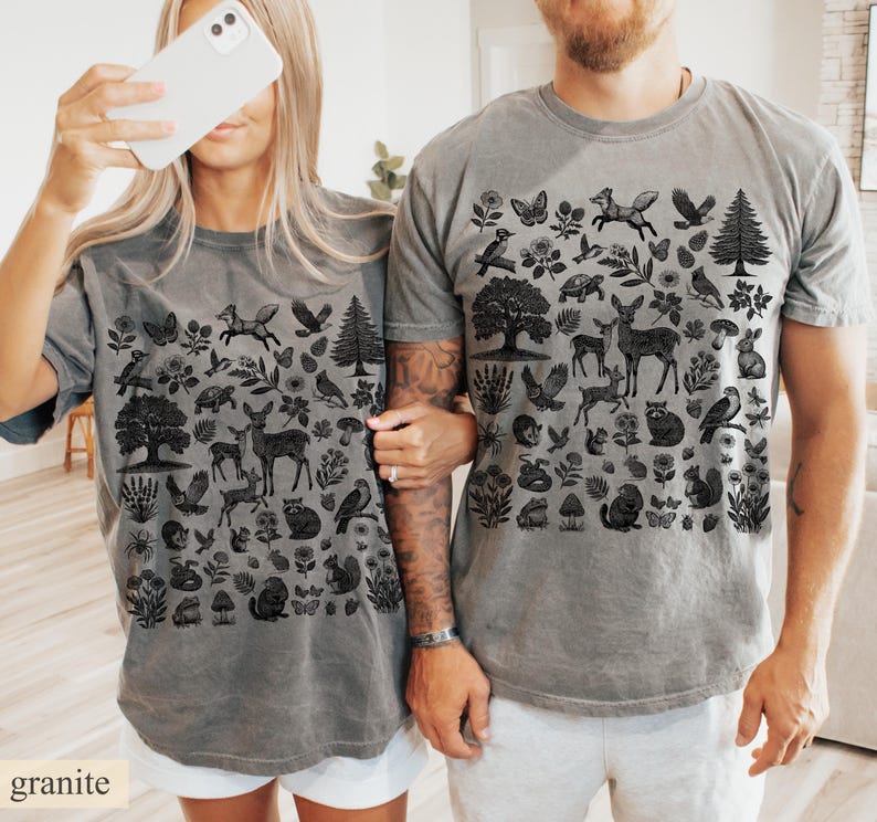 May include: Two gray t-shirts with a black nature-themed design. The design includes illustrations of trees, deer, birds, butterflies, and other woodland elements. The word "granite" is printed at the bottom left corner of the image.