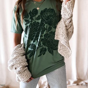 Puede incluir: Una camiseta de manga corta de color verde claro con un estampado gr&aacute;fico floral de color verde oscuro en la parte delantera. La prenda se combina con un c&aacute;rdigan de punto texturizado beige y vaqueros azul claro, creando un estilo casual.