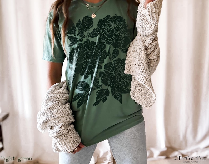 Puede incluir: Una camiseta de manga corta de color verde claro con un estampado gr&aacute;fico floral de color verde oscuro en la parte delantera. La prenda se combina con un c&aacute;rdigan de punto texturizado beige y vaqueros azul claro, creando un estilo casual.