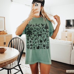 May include: A light green t-shirt featuring a black graphic of woodland creatures and plants. The design includes deer, foxes, birds, trees, and flowers. The shirt is a relaxed fit.