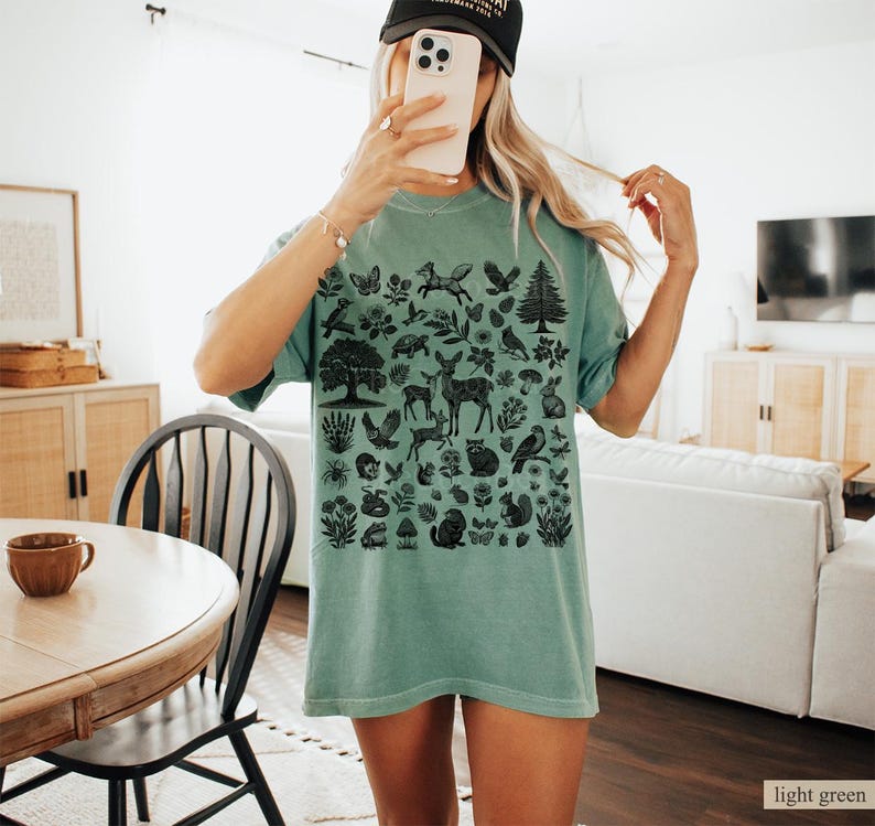 May include: A light green t-shirt featuring a black graphic of woodland creatures and plants. The design includes deer, foxes, birds, trees, and flowers. The shirt is a relaxed fit.