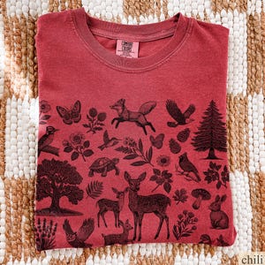 May include: A red t-shirt with a crew neck and a graphic print of various forest animals and plants. The design includes a fox, deer, birds, and trees. The shirt is made of a soft material and has a tag that says "Comfort Colors".