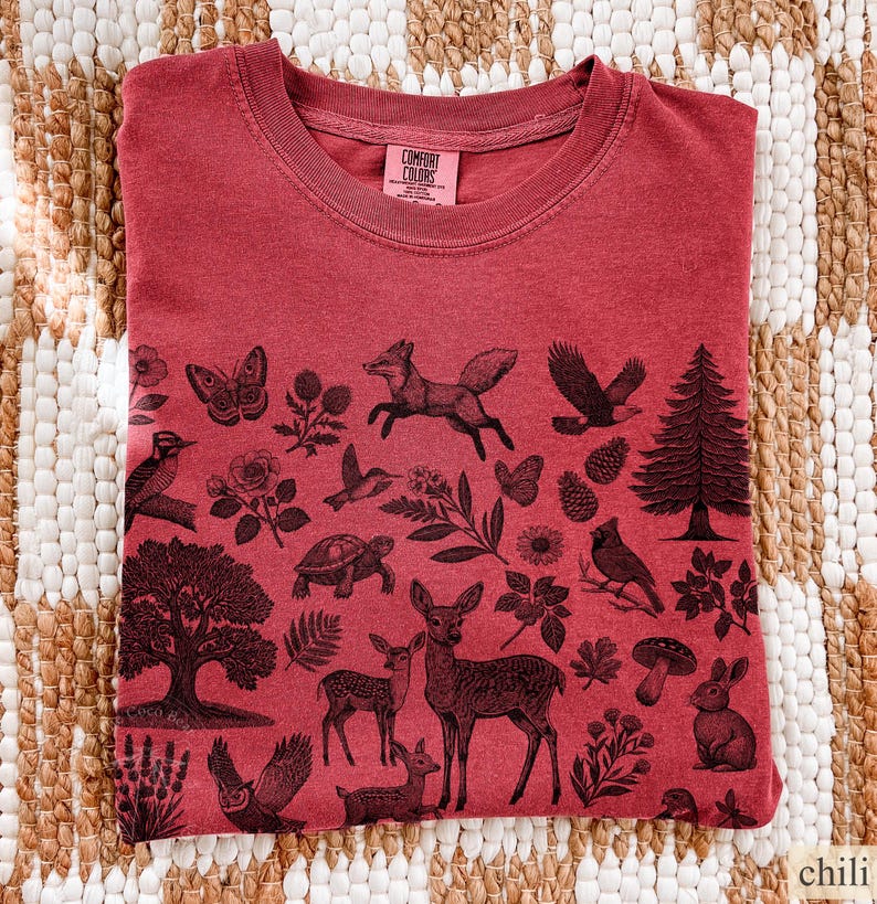 May include: A red t-shirt with a crew neck and a graphic print of various forest animals and plants. The design includes a fox, deer, birds, and trees. The shirt is made of a soft material and has a tag that says "Comfort Colors".