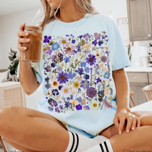 May include: A light blue t-shirt with a floral print featuring pressed flowers in shades of purple, blue, and yellow. The flowers are arranged in a random pattern and create a unique and eye-catching design.