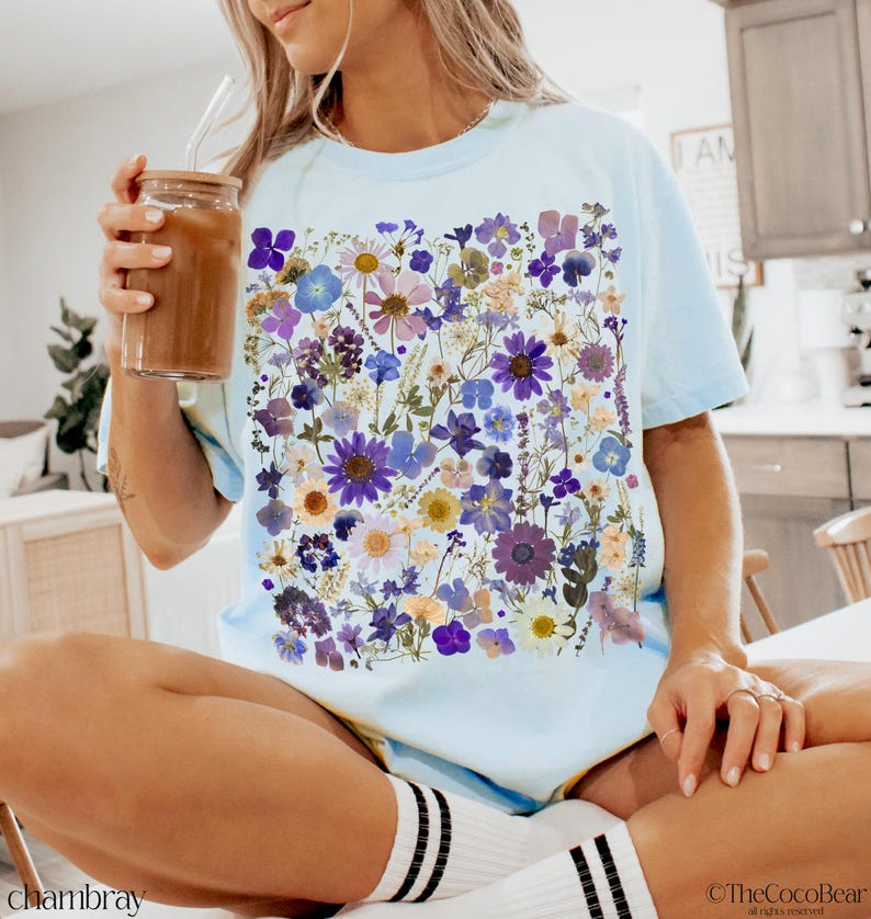 May include: A light blue t-shirt with a floral print featuring pressed flowers in shades of purple, blue, and yellow. The flowers are arranged in a random pattern and create a unique and eye-catching design.