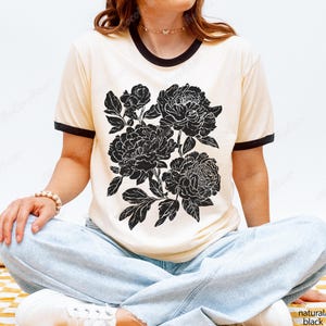 May include: Cream-colored ringer t-shirt with a black floral design. The graphic features a detailed illustration of peonies. The shirt has a black neckline and sleeve trim, and the text "natural/black" is visible.