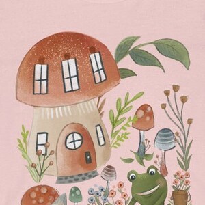 K&ouml;nnte beinhalten: Ein pinkfarbenes T-Shirt mit einer fantasievollen Illustration eines Pilzhauses, eines l&auml;chelnden Frosches und verschiedener Pilze in einer Waldlandschaft.