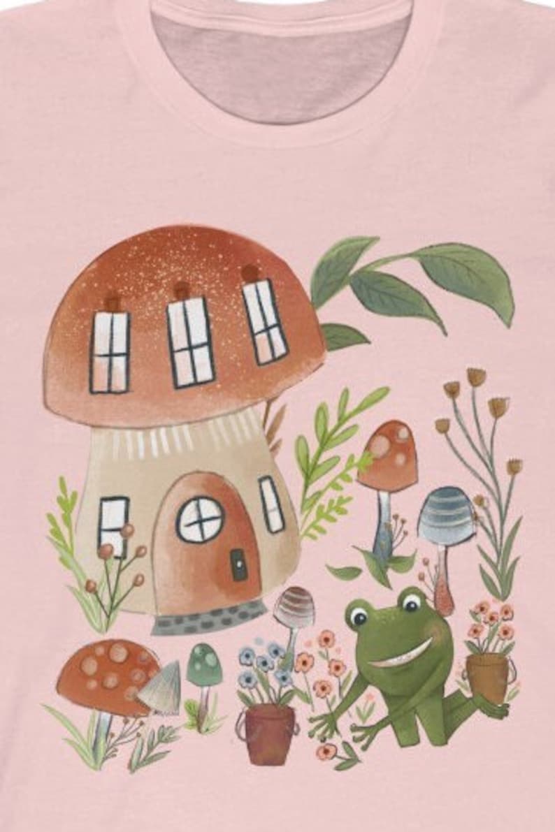 K&ouml;nnte beinhalten: Ein pinkfarbenes T-Shirt mit einer fantasievollen Illustration eines Pilzhauses, eines l&auml;chelnden Frosches und verschiedener Pilze in einer Waldlandschaft.