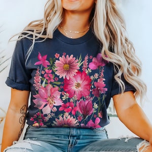 May include: Dark navy blue t-shirt with a colourful floral print in pink and magenta hues. The botanical design showcases a variety of flowers and leaves, forming a bright and artistic arrangement. The sleeves are rolled up.