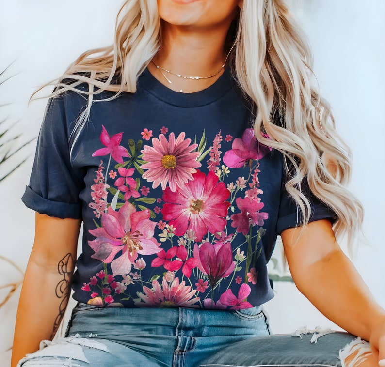 May include: Dark navy blue t-shirt with a colourful floral print in pink and magenta hues. The botanical design showcases a variety of flowers and leaves, forming a bright and artistic arrangement. The sleeves are rolled up.
