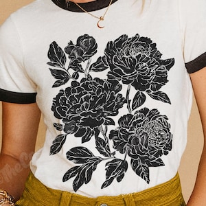 May include: White t-shirt with black trim featuring a large black and white floral design. The graphic includes detailed illustrations of peony flowers and leaves. The model is wearing gold jewelry and mustard-colored corduroy pants.