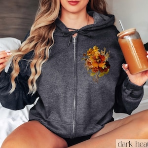 May include: A dark heather grey zippered hoodie with a floral print on the left chest. The print features a bouquet of yellow, orange, and brown flowers.