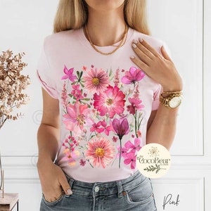 May include: A pale pink t-shirt with a watercolour floral print in pink and magenta hues. The design showcases diverse flowers and leaves. The word "Pink" is visible at the bottom right.