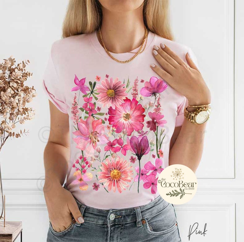 May include: A pale pink t-shirt with a watercolour floral print in pink and magenta hues. The design showcases diverse flowers and leaves. The word "Pink" is visible at the bottom right.