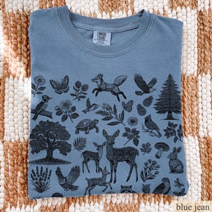May include: Blue jean colored t-shirt with a nature-themed graphic. The design features various animals and plants, including a fox, deer, owl, and pine tree. The shirt is folded and displayed on a woven surface.