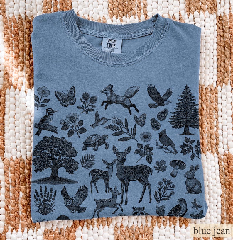 May include: Blue jean colored t-shirt with a nature-themed graphic. The design features various animals and plants, including a fox, deer, owl, and pine tree. The shirt is folded and displayed on a woven surface.