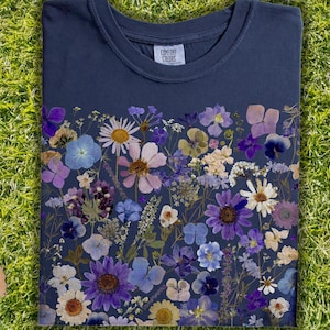 May include: A navy blue t-shirt with a floral design made from pressed flowers. The flowers are in shades of purple, blue, and white. The shirt has a crew neck and short sleeves. The text "Comfort Colors" is printed on the tag.
