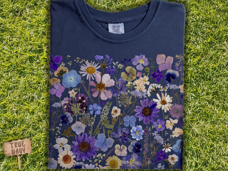May include: A navy blue t-shirt with a floral design made from pressed flowers. The flowers are in shades of purple, blue, and white. The shirt has a crew neck and short sleeves. The text "Comfort Colors" is printed on the tag.