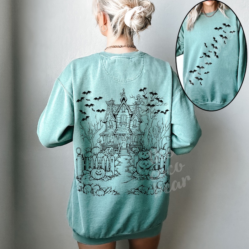 Op de afbeelding: Een tealblauw sweatshirt met een zwart-witte illustratie van een spookhuis met vleermuizen en pompoenen. Het sweatshirt heeft een ronde hals en lange mouwen.