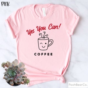 Może przedstawiać: R&oacute;żowa koszulka z napisem "Yes You Can!" w czerwonym skrypcie nad grafiką mrugającej filiżanki kawy. Poniżej grafiki słowo "COFFEE" jest wydrukowane czarnymi literami. Koszulka jest w kolorze jasnor&oacute;żowym.