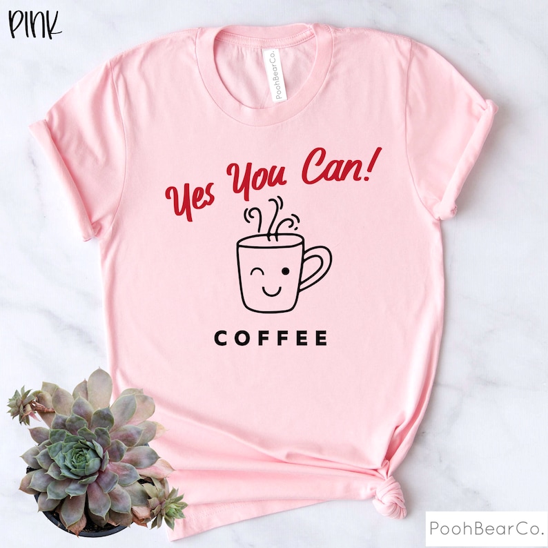 Może przedstawiać: R&oacute;żowa koszulka z napisem "Yes You Can!" w czerwonym skrypcie nad grafiką mrugającej filiżanki kawy. Poniżej grafiki słowo "COFFEE" jest wydrukowane czarnymi literami. Koszulka jest w kolorze jasnor&oacute;żowym.