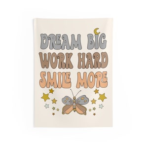 May include: A beige tapestry with the text "Dream Big Work Hard Smile More" in a retro font. A butterfly with orange, gray, and brown wings is centered below the text. The butterfly is surrounded by gold and white stars.