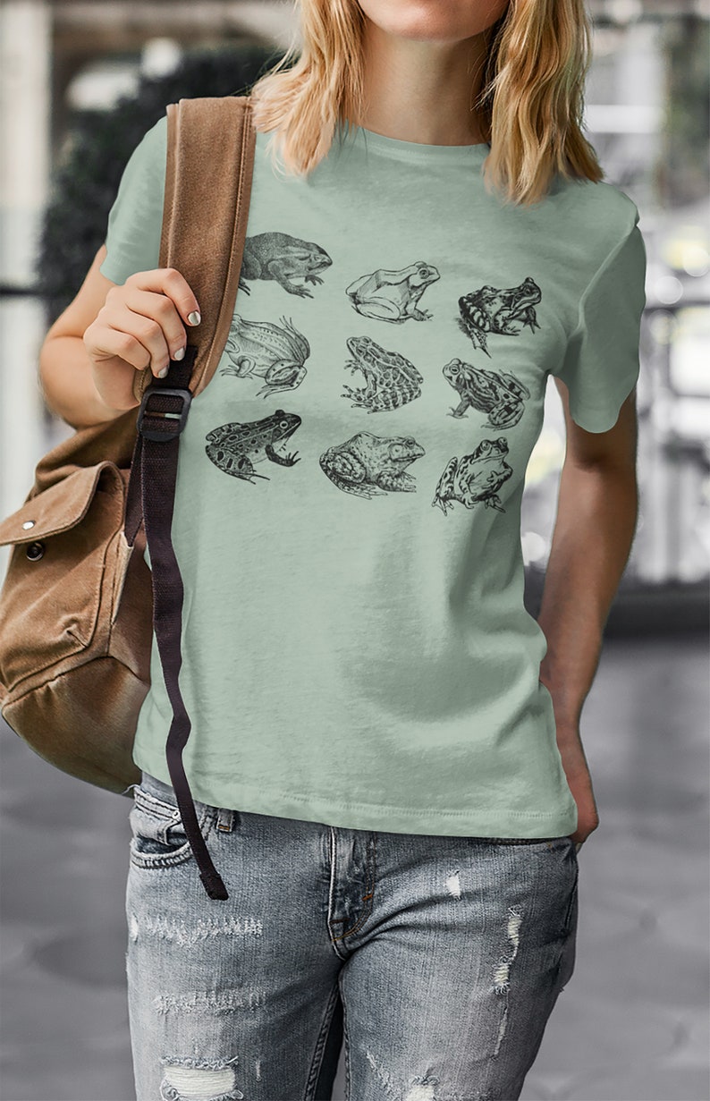 Puede incluir: Camiseta verde claro con ilustraciones detalladas en blanco y negro de varias especies de ranas. La camiseta se combina con vaqueros azules. Se ve un bolso de hombro marr&oacute;n.