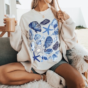 Puede incluir: Una sudadera blanca con un estampado de conchas marinas azules y blancas. La sudadera la lleva puesta una persona sentada en un sof&aacute; blanco.
