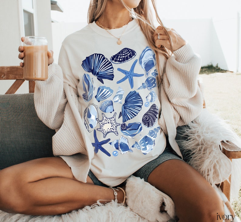 Puede incluir: Una sudadera blanca con un estampado de conchas marinas azules y blancas. La sudadera la lleva puesta una persona sentada en un sof&aacute; blanco.