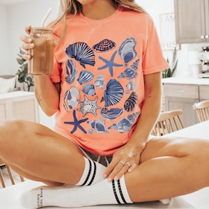 Puede incluir: Una mujer lleva una camiseta de color coral con un estampado de conchas marinas azules y blancas. Est&aacute; sentada en una encimera blanca con las piernas cruzadas y sosteniendo un vaso de caf&eacute; helado.
