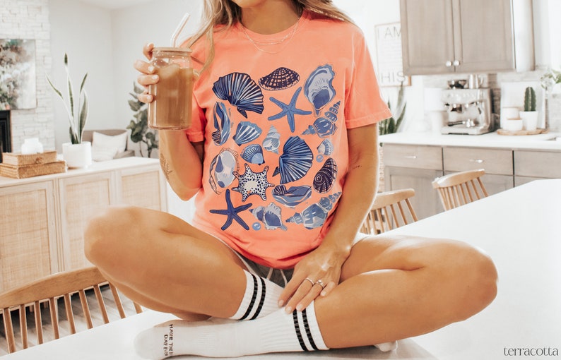 Puede incluir: Una mujer lleva una camiseta de color coral con un estampado de conchas marinas azules y blancas. Est&aacute; sentada en una encimera blanca con las piernas cruzadas y sosteniendo un vaso de caf&eacute; helado.