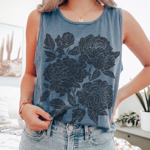 May include: A blue sleeveless tank top featuring a large, dark gray floral graphic print on the front. The design displays detailed illustrations of blooming flowers and leaves. The garment has a round neckline and is paired with light blue denim jeans.