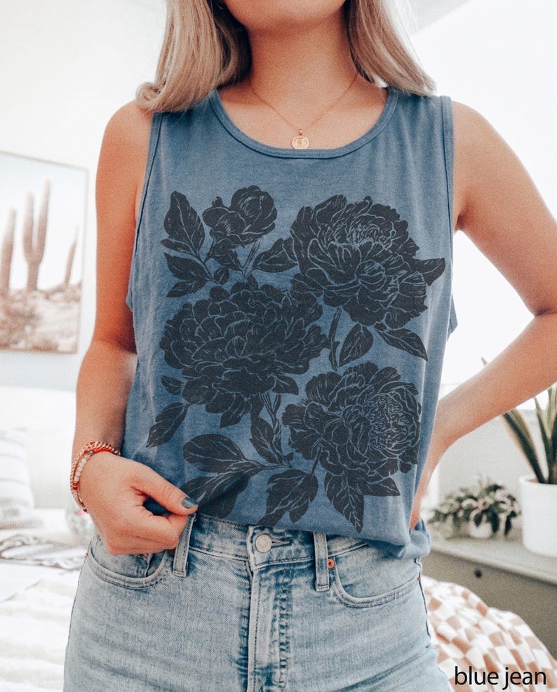 May include: A blue sleeveless tank top featuring a large, dark gray floral graphic print on the front. The design displays detailed illustrations of blooming flowers and leaves. The garment has a round neckline and is paired with light blue denim jeans.
