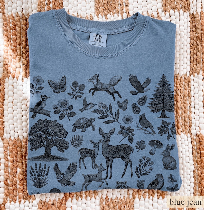 May include: Blue jean colored t-shirt with a nature-themed graphic. The design features various animals and plants, including a fox, deer, owl, and pine tree. The shirt is folded and displayed on a woven surface.