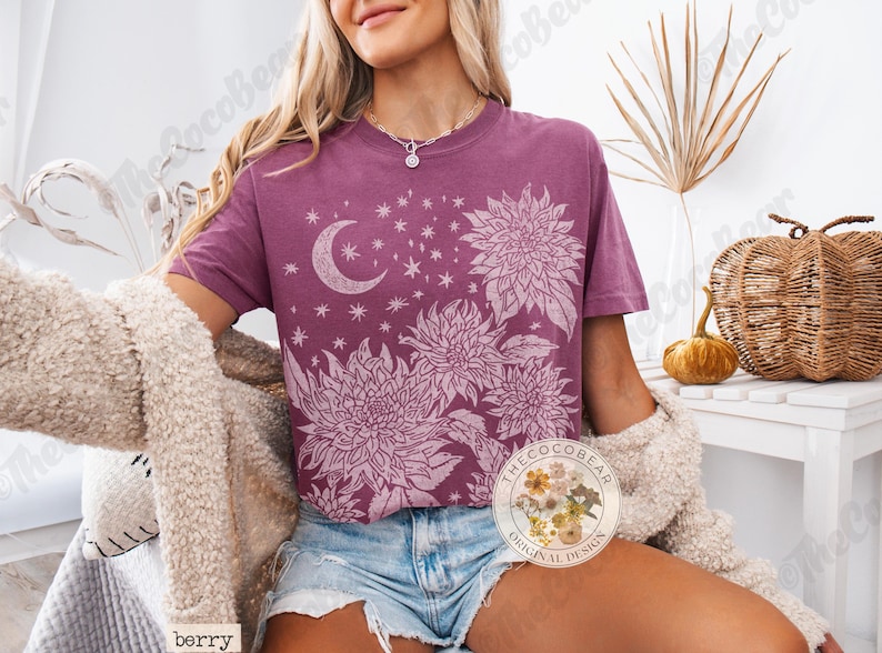 Pode incluir: Camiseta cor de berry com um design floral e celestial. O gr&aacute;fico inclui uma lua crescente, estrelas e flores grandes. A camisa tem gola redonda e caimento solto. Design original CocoBear.