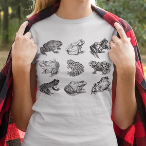 Puede incluir: Camiseta blanca con una ilustraci&oacute;n en blanco y negro de nueve especies de ranas. Las ranas est&aacute;n representadas en varias poses y texturas, dispuestas en tres filas de tres. La camiseta est&aacute; hecha de un material suave.