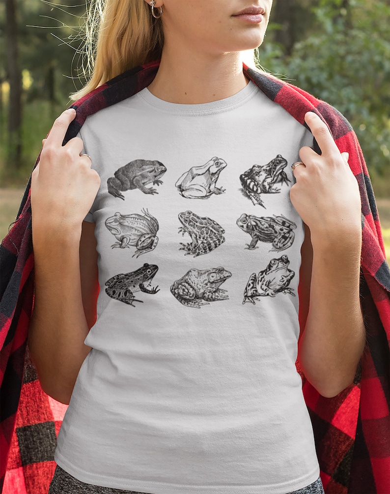 Puede incluir: Camiseta blanca con una ilustraci&oacute;n en blanco y negro de nueve especies de ranas. Las ranas est&aacute;n representadas en varias poses y texturas, dispuestas en tres filas de tres. La camiseta est&aacute; hecha de un material suave.