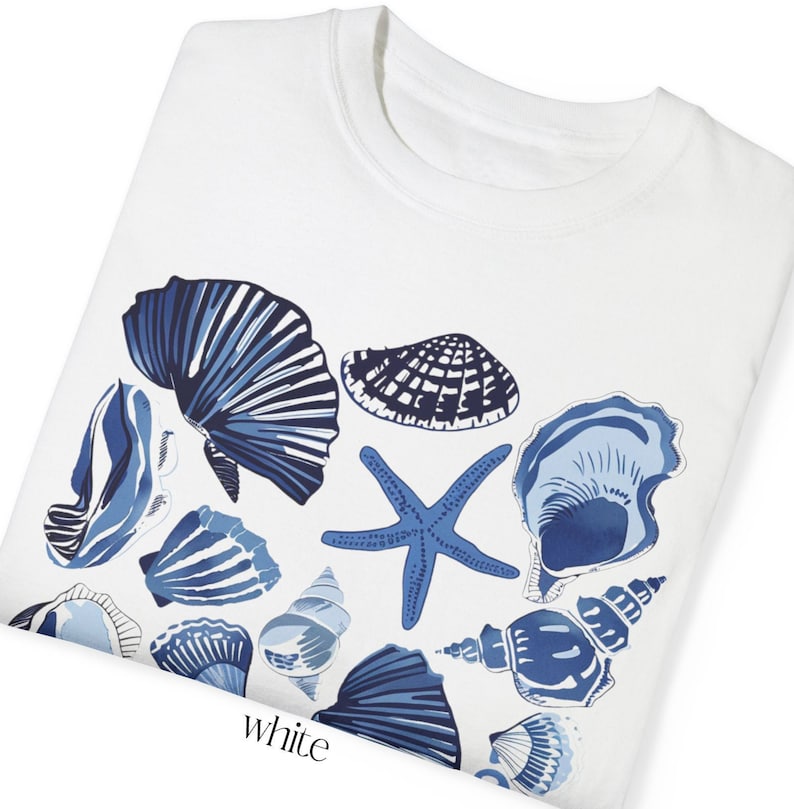 Puede incluir: Camiseta blanca con un dise&ntilde;o de conchas marinas azul y blanco. El dise&ntilde;o incluye varias conchas, una estrella de mar y algunas conchas m&aacute;s peque&ntilde;as.