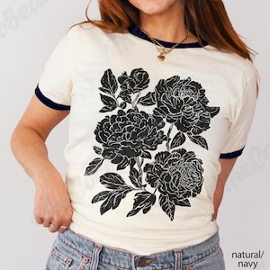May include: Cream-colored t-shirt with navy blue trim featuring a large black floral design. The shirt has short sleeves and a round neckline. The text "natural/navy" is visible in the bottom right corner.