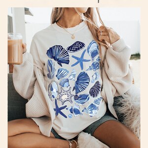 Puede incluir: Una sudadera blanca con un dise&ntilde;o de conchas marinas y estrellas de mar azules y blancas. El texto "thecocobear.etsy.com" est&aacute; impreso en la parte inferior de la imagen.