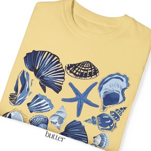 Puede incluir: Una camiseta amarilla con un dise&ntilde;o de conchas marinas azules y blancas. El dise&ntilde;o incluye una estrella de mar, varias conchas y la palabra "butter" impresa en la parte inferior.