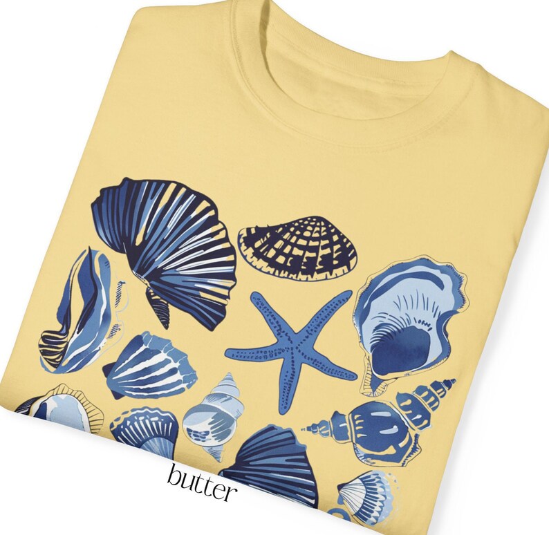 Puede incluir: Una camiseta amarilla con un dise&ntilde;o de conchas marinas azules y blancas. El dise&ntilde;o incluye una estrella de mar, varias conchas y la palabra "butter" impresa en la parte inferior.