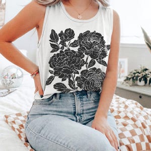 May include: A white sleeveless tank top featuring a large black graphic print of blooming peonies with detailed petals and leaves. The garment has a round neckline and is paired with light blue denim jeans. The design is centered on the front.