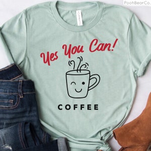 Może przedstawiać: Jasnozielona koszulka z czerwonym napisem "Yes You Can!" nad grafiką mrugającego kubka do kawy. Słowo "COFFEE" jest wydrukowane poniżej na czarno. Koszulka jest prezentowana z niebieskimi dżinsami i brązowym butem.