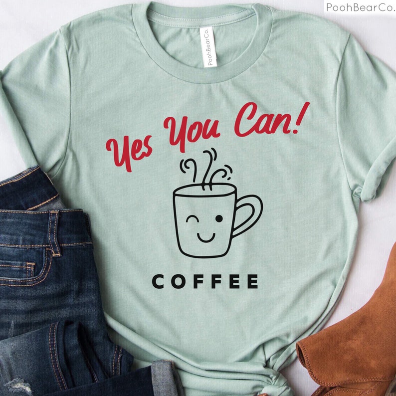 Może przedstawiać: Jasnozielona koszulka z czerwonym napisem "Yes You Can!" nad grafiką mrugającego kubka do kawy. Słowo "COFFEE" jest wydrukowane poniżej na czarno. Koszulka jest prezentowana z niebieskimi dżinsami i brązowym butem.