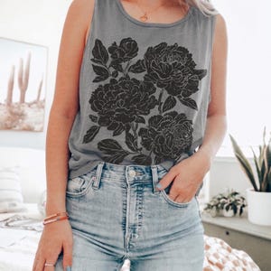 May include: A grey sleeveless tank top featuring a large black graphic print of three blooming peonies with detailed leaves and petals. The shirt has a round neckline and is paired with light blue denim jeans that have a classic button fly closure.